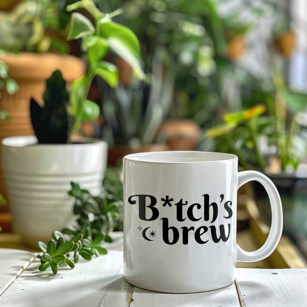 A white ceramic coffee mug with the phrase B*tch's Brew on the side. Plants in the background.