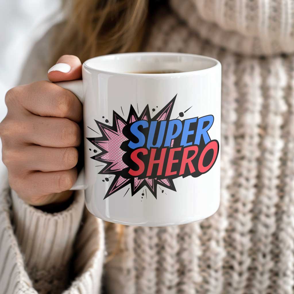 A woman holding a white ceramic coffee mug with the phrase 'SUPER SHERO' in bold, colorful comic book-style lettering, featuring a burst effect in the background.